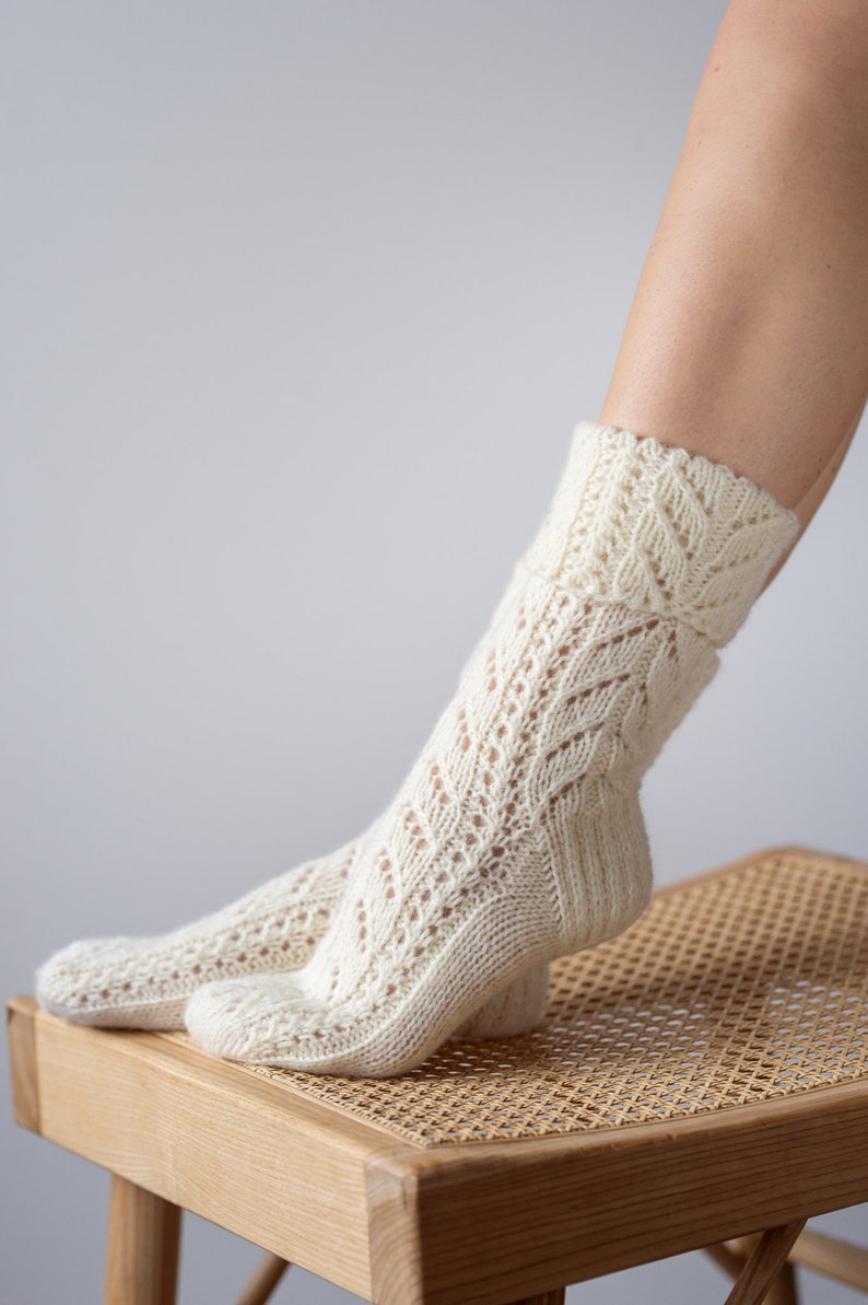 May include: A pair of white knitted socks with a lacy pattern. The socks are on a wooden stool with a woven seat.
