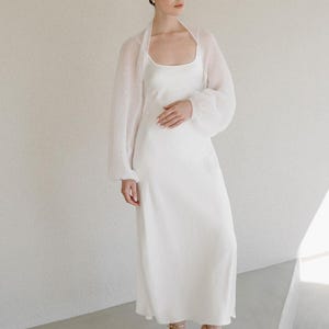 May include: A floor-length white slip dress with a scoop neckline and a matching sheer, long-sleeved white knit bolero cardigan. The outfit is paired with gold strappy high-heeled sandals featuring multiple thin straps across the front of the foot.