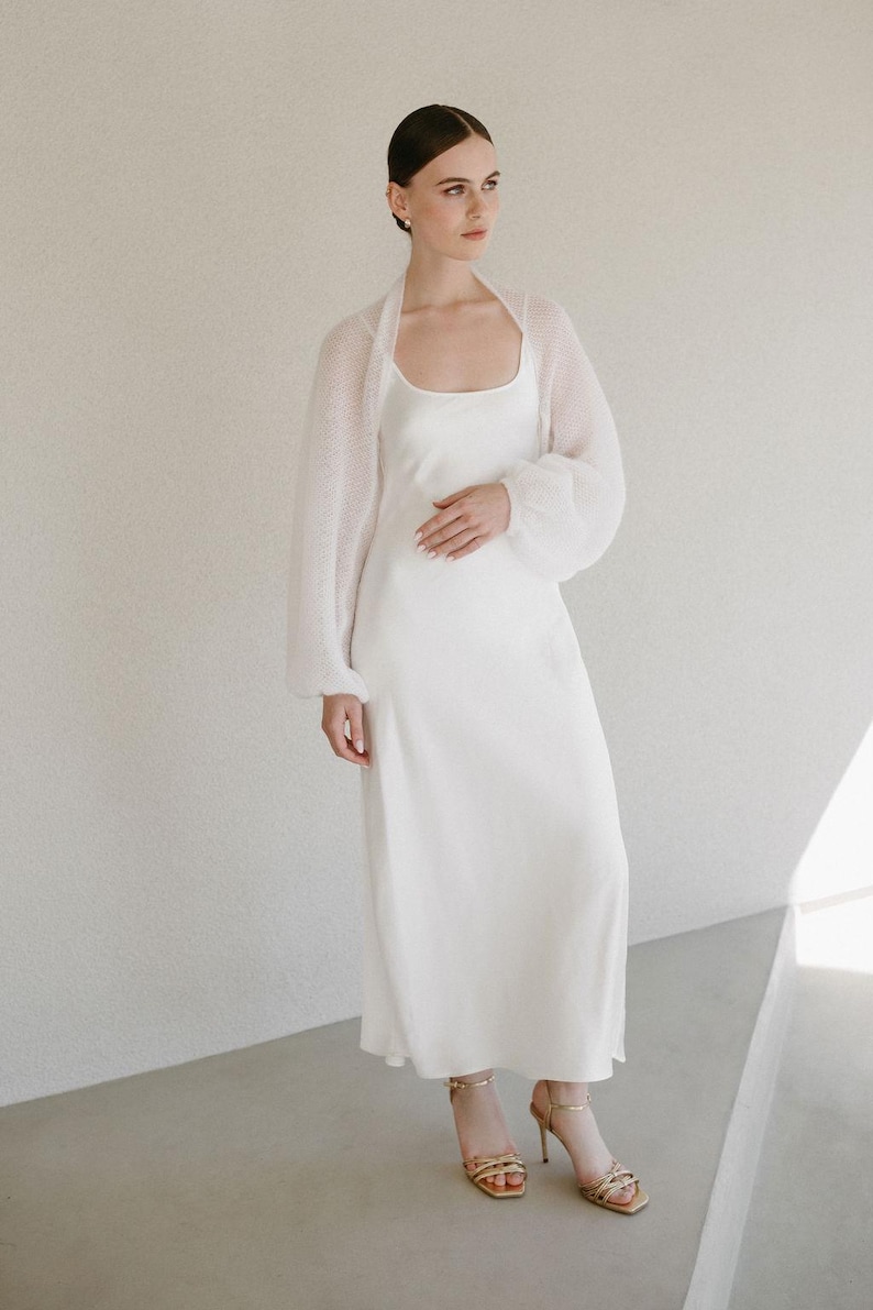 May include: A floor-length white slip dress with a scoop neckline and a matching sheer, long-sleeved white knit bolero cardigan. The outfit is paired with gold strappy high-heeled sandals featuring multiple thin straps across the front of the foot.