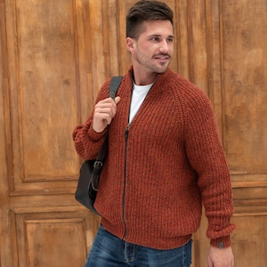 May include: A man wearing a rust-colored knitted zip-up cardigan with a white t-shirt underneath. He is carrying a black leather bag over his shoulder and wearing blue jeans.