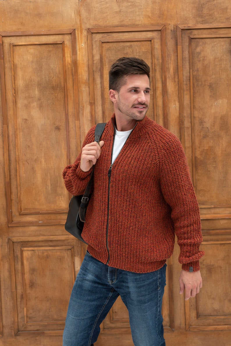 May include: A man wearing a rust-colored knitted zip-up cardigan with a white t-shirt underneath. He is carrying a black leather bag over his shoulder and wearing blue jeans.