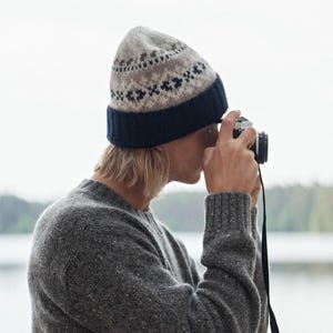 May include: A person wearing a gray and brown patterned knit beanie and a gray sweater is holding a vintage camera and looking through the viewfinder.
