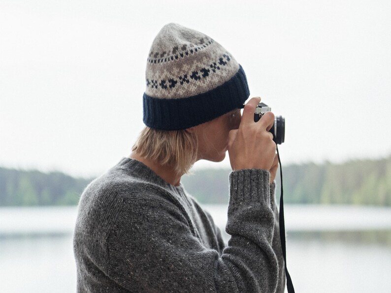 May include: A person wearing a gray and brown patterned knit beanie and a gray sweater is holding a vintage camera and looking through the viewfinder.