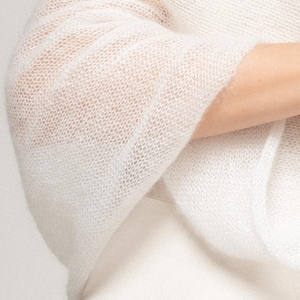 May include: A white, semi-sheer, knitted shawl with a loose, open-weave texture. The fabric appears lightweight and airy, featuring a fine, delicate knit pattern that drapes softly. The garment is solid white and has a soft, slightly fuzzy appearance.