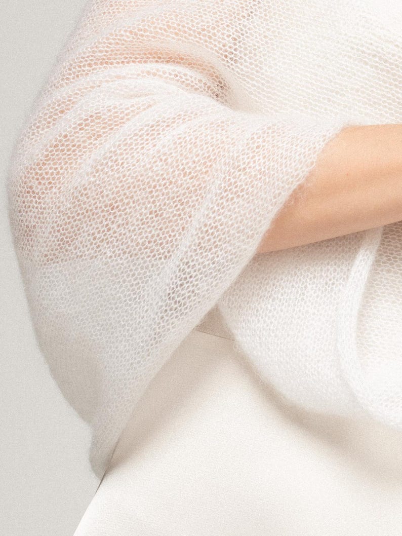 May include: A white, semi-sheer, knitted shawl with a loose, open-weave texture. The fabric appears lightweight and airy, featuring a fine, delicate knit pattern that drapes softly. The garment is solid white and has a soft, slightly fuzzy appearance.