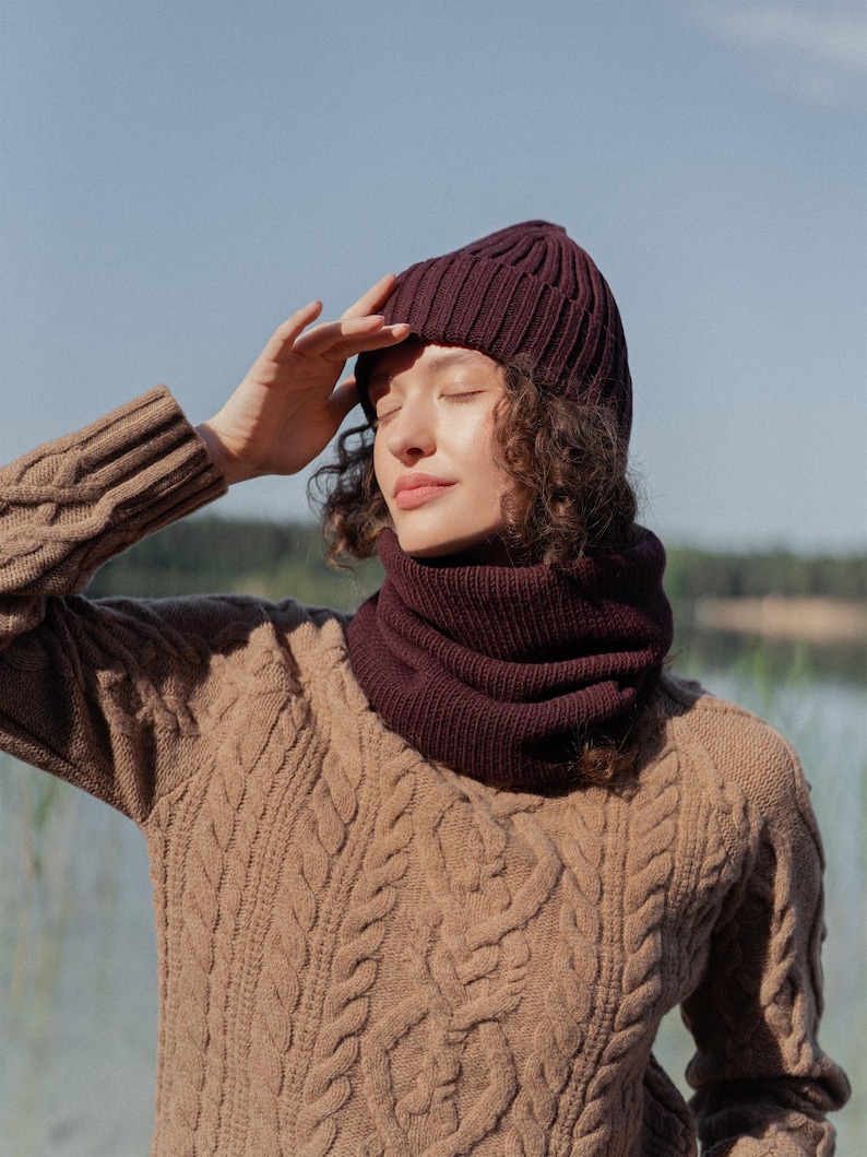 May include: A dark burgundy ribbed knit beanie and a matching dark burgundy ribbed knit neck warmer. The beanie features a folded cuff design, and both items are crafted from a thick, textured yarn, providing a cohesive and warm winter accessory set.