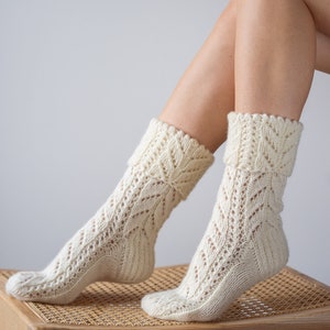 May include: A pair of white knitted socks with a lacy pattern. The socks are worn by a person sitting on a wooden stool with a woven seat.