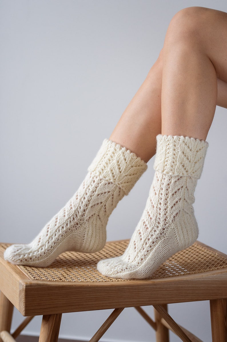 May include: A pair of white knitted socks with a lacy pattern. The socks are worn by a person sitting on a wooden stool with a woven seat.