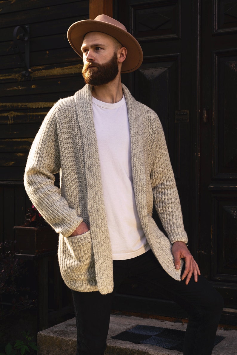 May include: A man wearing a light brown fedora hat, a white t-shirt, and a light gray cardigan with a shawl collar. He is standing in front of a dark wooden door.