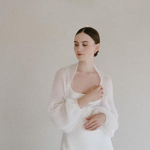May include: A white knitted bolero with long, voluminous sleeves and a soft, semi-sheer texture. The garment features a simple, open-front design with a rounded neckline, crafted from a lightweight, airy yarn that drapes gently over the shoulders.