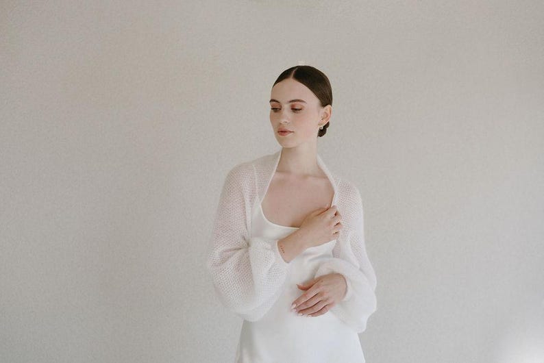 May include: A white knitted bolero with long, voluminous sleeves and a soft, semi-sheer texture. The garment features a simple, open-front design with a rounded neckline, crafted from a lightweight, airy yarn that drapes gently over the shoulders.