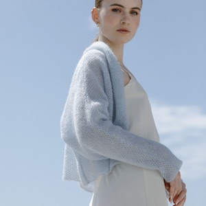 May include: A light blue, open-front knit cardigan with a textured, airy stitch pattern, worn over a simple, sleeveless cream-colored slip dress. The cardigan features long sleeves and a relaxed, cropped silhouette that drapes softly over the shoulders.