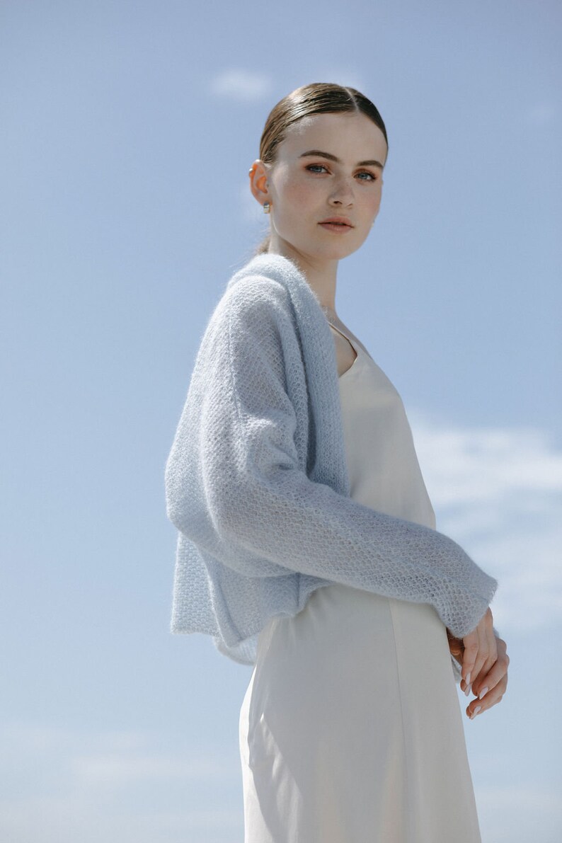 May include: A light blue, open-front knit cardigan with a textured, airy stitch pattern, worn over a simple, sleeveless cream-colored slip dress. The cardigan features long sleeves and a relaxed, cropped silhouette that drapes softly over the shoulders.