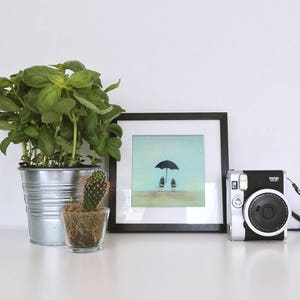 May include: A framed illustration of two figures standing under a black umbrella on a light blue background. A potted cactus and a potted basil plant are in the foreground. A silver instant camera with a black strap is on a white surface.