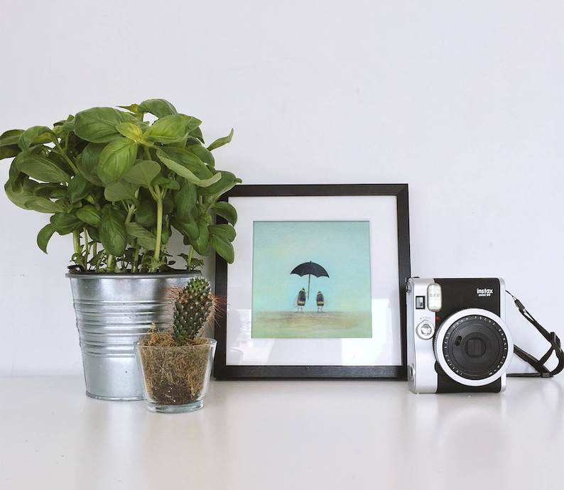 May include: A framed illustration of two figures standing under a black umbrella on a light blue background. A potted cactus and a potted basil plant are in the foreground. A silver instant camera with a black strap is on a white surface.