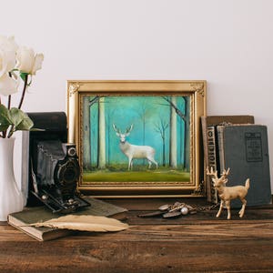 Pode incluir: Uma pintura emoldurada de um veado branco em p&eacute; em uma floresta com um fundo azul e verde. A pintura est&aacute; sobre uma mesa de madeira com um vaso de rosas brancas, uma c&acirc;mera vintage, livros e uma figura de veado dourada.