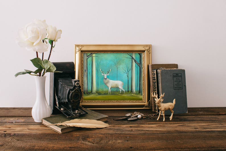 Pode incluir: Uma pintura emoldurada de um veado branco em p&eacute; em uma floresta com um fundo azul e verde. A pintura est&aacute; sobre uma mesa de madeira com um vaso de rosas brancas, uma c&acirc;mera vintage, livros e uma figura de veado dourada.