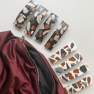 May include: Set of six colorful hair clips with a terrazzo pattern. The clips are made of resin and have a curved shape. They are arranged in two rows on a white surface.