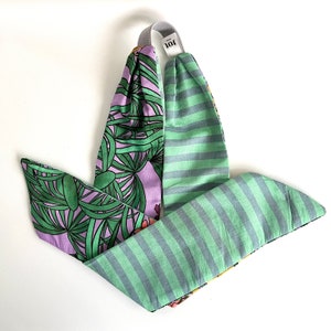 Op de afbeelding: Een groen en paars hoofdband met een tropische bladprint aan de ene kant en een groen-wit gestreept patroon aan de andere kant. Het hoofdband is in een knoop geknoopt en heeft een klein label met de tekst "JOI".