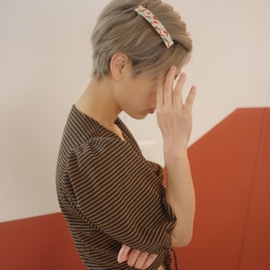May include: A person with short, gray hair wearing a brown and black striped shirt with a tie at the waist. The person is wearing a patterned hair clip.