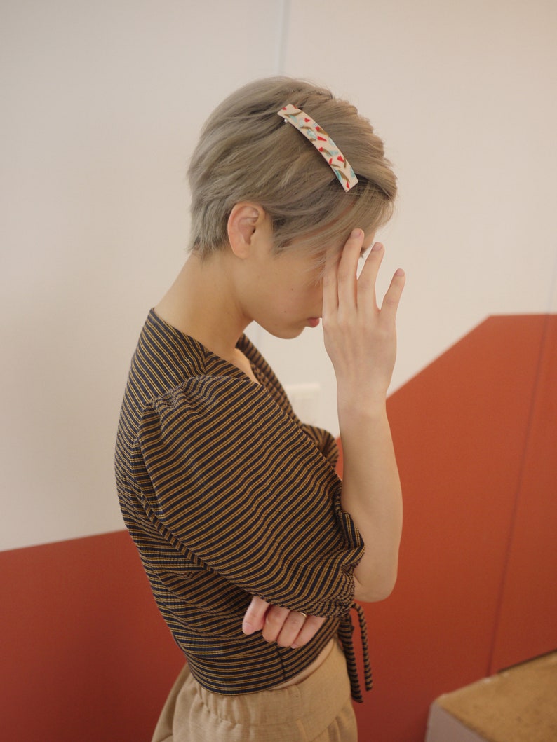 May include: A person with short, gray hair wearing a brown and black striped shirt with a tie at the waist. The person is wearing a patterned hair clip.