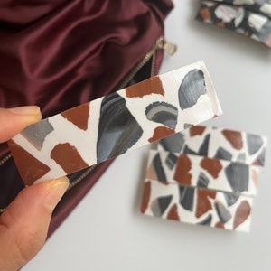 May include: A white rectangular hair clip with a terrazzo pattern in shades of brown, black, and gray.