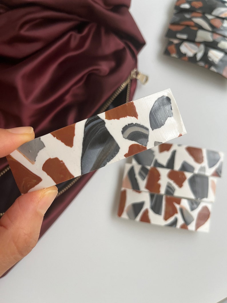 May include: A white rectangular hair clip with a terrazzo pattern in shades of brown, black, and gray.