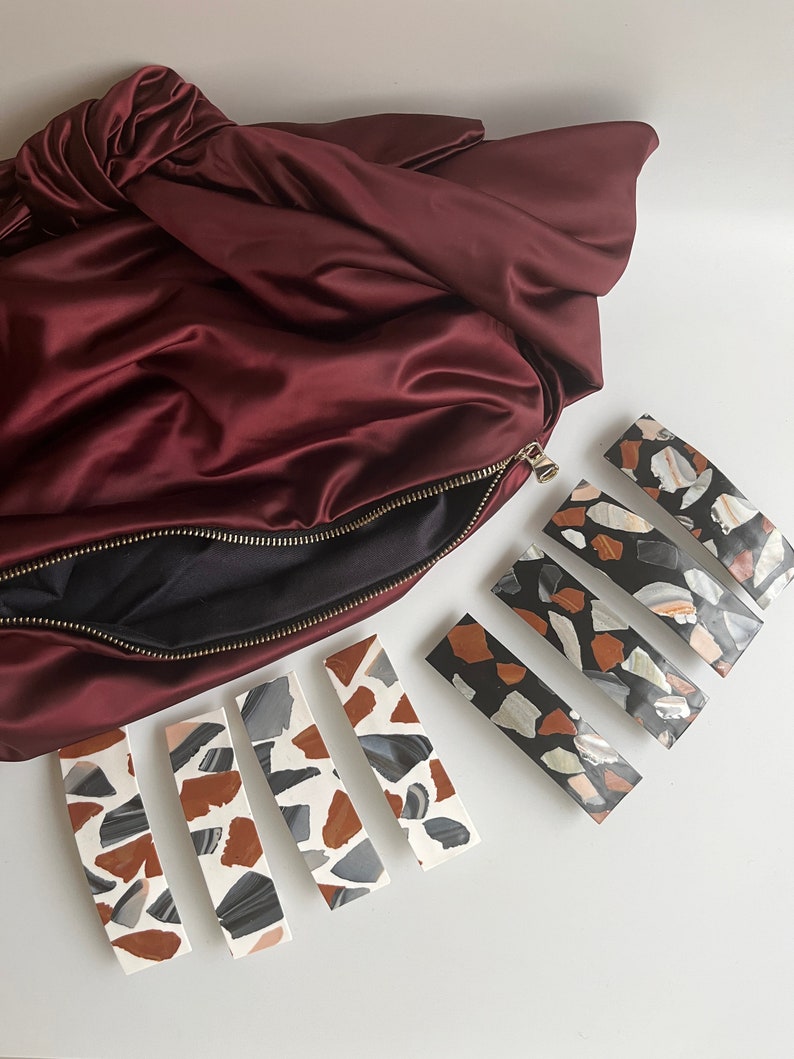 May include: Five hair clips with a terrazzo pattern. The clips are in two different color schemes: black and white, and brown, white, and gray. The clips are arranged in a row on a white surface.