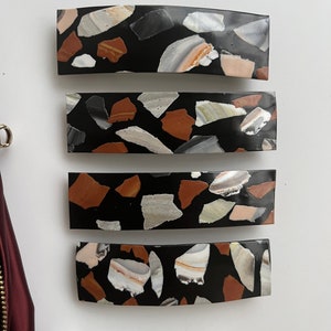 May include: Four black hair clips with a terrazzo pattern. The clips are curved and have a black background with white, brown, and beige flecks.