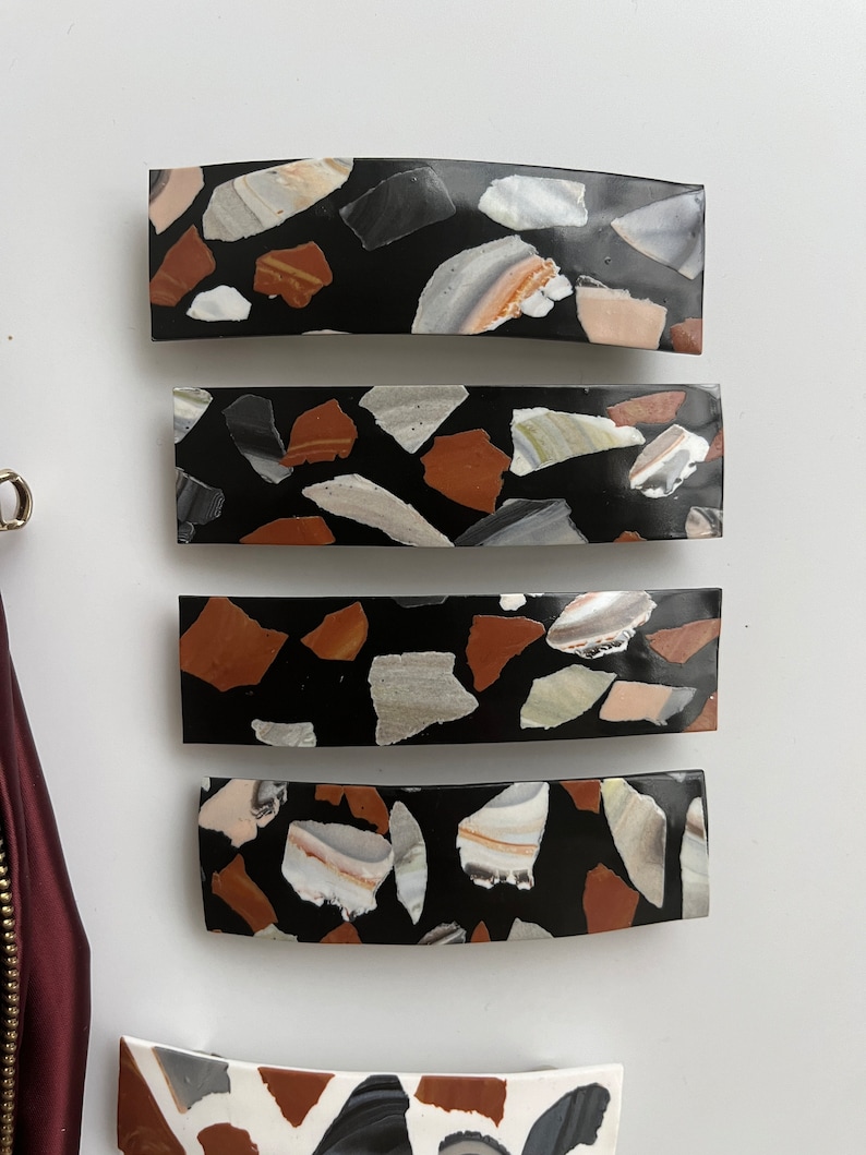 May include: Four black hair clips with a terrazzo pattern. The clips are curved and have a black background with white, brown, and beige flecks.