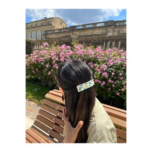 May include: A woman with long dark hair is wearing a white and green patterned hair clip. She is sitting on a wooden bench in front of a pink flower bush.