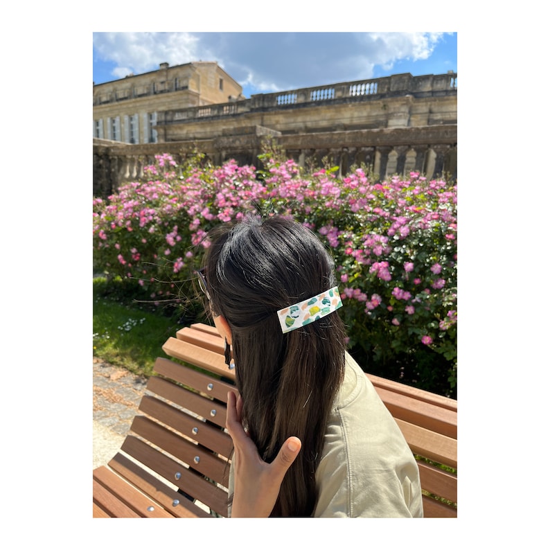 May include: A woman with long dark hair is wearing a white and green patterned hair clip. She is sitting on a wooden bench in front of a pink flower bush.