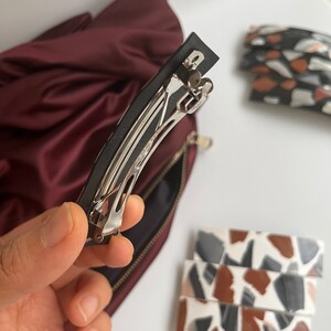 May include: A silver metal hair clip with a black plastic base. The clip is open and ready to be used to hold hair in place.