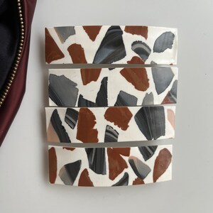 May include: Four white hair clips with a terrazzo pattern. The pattern features brown, gray, and black pieces.