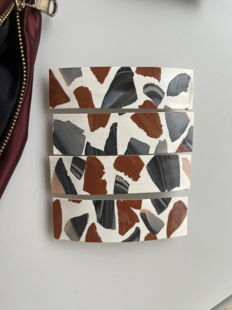 May include: Four white hair clips with a terrazzo pattern. The pattern features brown, gray, and black pieces.