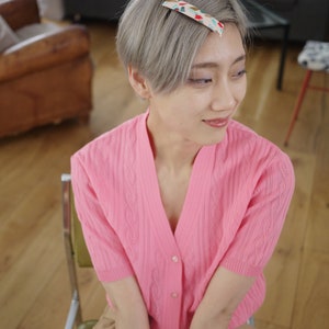 May include: A woman with short, silver hair wearing a pink cardigan with a cable knit pattern and a brown skirt. She is wearing a patterned hair clip in her hair.