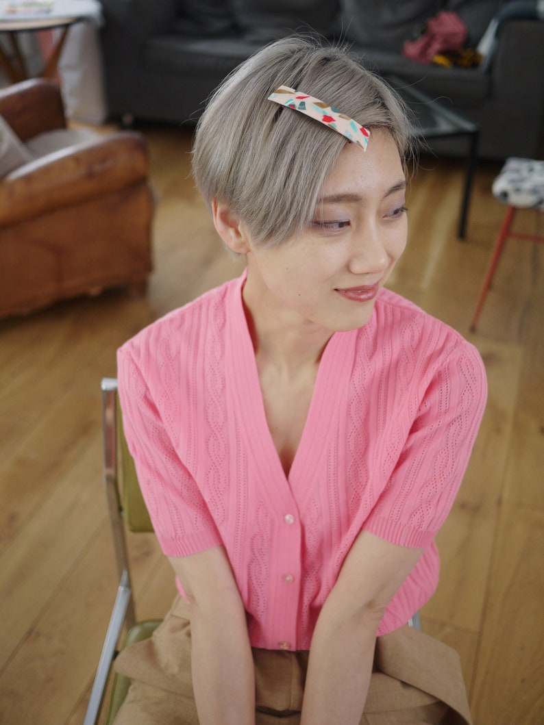 May include: A woman with short, silver hair wearing a pink cardigan with a cable knit pattern and a brown skirt. She is wearing a patterned hair clip in her hair.