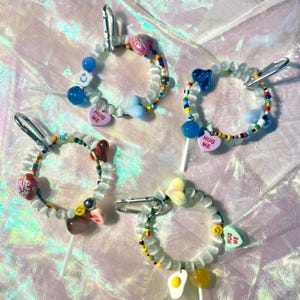 May include: Four colourful beaded bracelets with charms, each featuring a different candy lollipop with a heart-shaped charm that says "Hug Me".