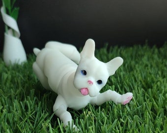 May include: A white, small, plastic figurine of a dog with blue eyes and a pink tongue sticking out. The dog is lying on its side with its head turned to the side. The figurine is on a green, artificial grass surface.