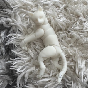 May include: A white, articulated cat-shaped ball-jointed doll featuring a smooth, matte finish. The figure has a feline head with pointed ears, a segmented torso, movable limbs, and a long, jointed tail, allowing for various poses.