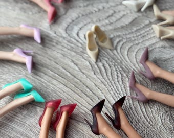 Puede incluir: Una colección de piernas de muñeca con zapatos de tacón alto en varios colores, incluyendo turquesa, blanco, beige, lavanda, rojo y marrón oscuro, dispuestos en un patrón circular con un par de tacones beige en el centro.
