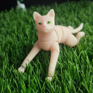 May include: A white articulated doll cat with green eyes lying on green grass.