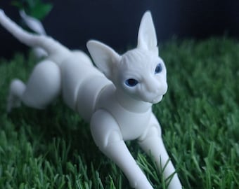 May include: A white, hairless cat doll with blue eyes, posed in a playful, crouching position on a green artificial grass surface.