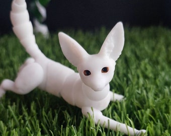 May include: A white, articulated, miniature fox figurine with orange eyes. The figurine is posed in a playful, running position on a green artificial grass surface.