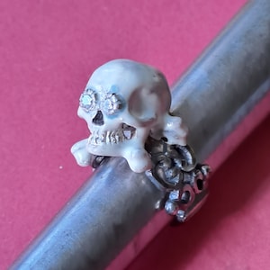 Op de afbeelding: Een schedelring met een wit schedel- en kruisbotontwerp. De schedel heeft sprankelende accenten voor de ogen en is gezet op een zilveren band. De ring wordt getoond op een zilveren staaf tegen een roze achtergrond.