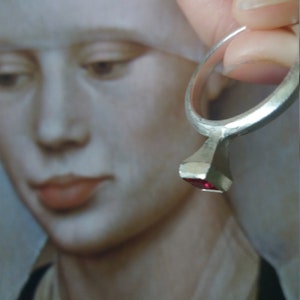 Op de afbeelding: Een zilveren ring met een vierkante rode edelsteen. De ring wordt vastgehouden door een hand voor een schilderij van een vrouw.