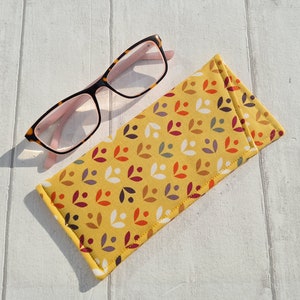 May include: A yellow fabric eyeglasses case with a pattern of small white, orange, red, and purple leaves. The case is closed and lying on a white wooden surface. A pair of pink and tortoise shell eyeglasses is lying next to the case.
