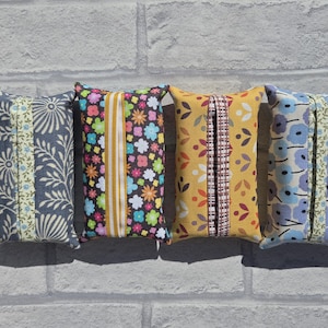May include: Four rectangular fabric tissue box covers arranged in a row. The first is grey with white floral patterns, the second is black with colourful flowers, the third is yellow with orange and red shapes, and the fourth is light blue with purple flowers.