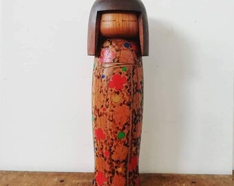 May include: A wooden Japanese kokeshi doll with a brown head and a colorful floral pattern on the body. The doll is standing on a wooden surface.
