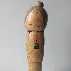 Könnte beinhalten: Japanische Kokeshi-Puppe aus Holz mit hellbraunem Finish. Die Puppe hat ein bemaltes Gesicht mit geschlossenen Augen und einem kleinen roten Mund. Der Körper ist mit goldenen Blumenmustern und schwarzen und roten Akzenten verziert.